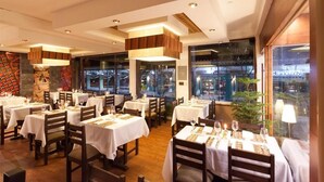 A restaurant with multiple tables, each set with white tablecloths and chairs, and large windows offering a view of the city. - Inti Punku Alameda Inn (Machu Picchu)