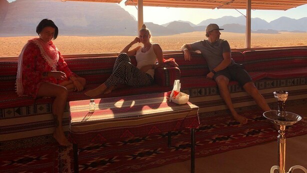 A room with a large window overlooking a desert landscape. There are three people seated on a patterned sofa. - camp Nights stars (Wadi Rum)