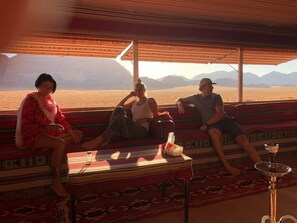 A room with a large window overlooking a desert landscape. There are three people seated on a patterned sofa. - camp Nights stars (Wadi Rum)