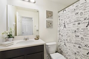A bathroom with a white sink, dark cabinets, a toilet, and a mirror. There are decorative items on the counter and a shower curtain with text. - Villa Garcia - West Haven (Davenport)
