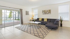 A modern living room with a grey sofa, a white coffee table, and a patterned rug. There is a sliding glass door leading to a pool area.