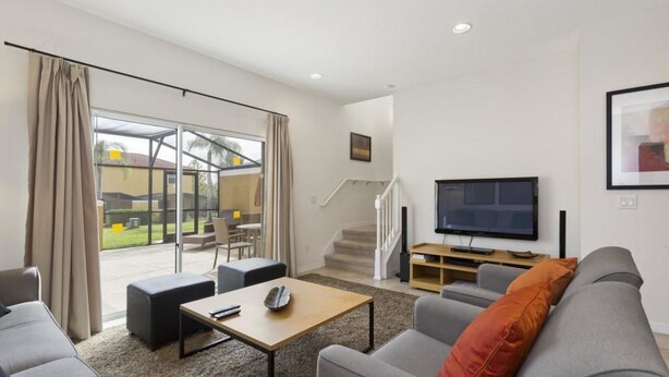 A modern living room with a grey sofa, a wooden coffee table, and a flat-screen TV. There is a staircase leading to an outdoor area with a glass door. - Villa Magical - Encantada Resort (Kissimmee)