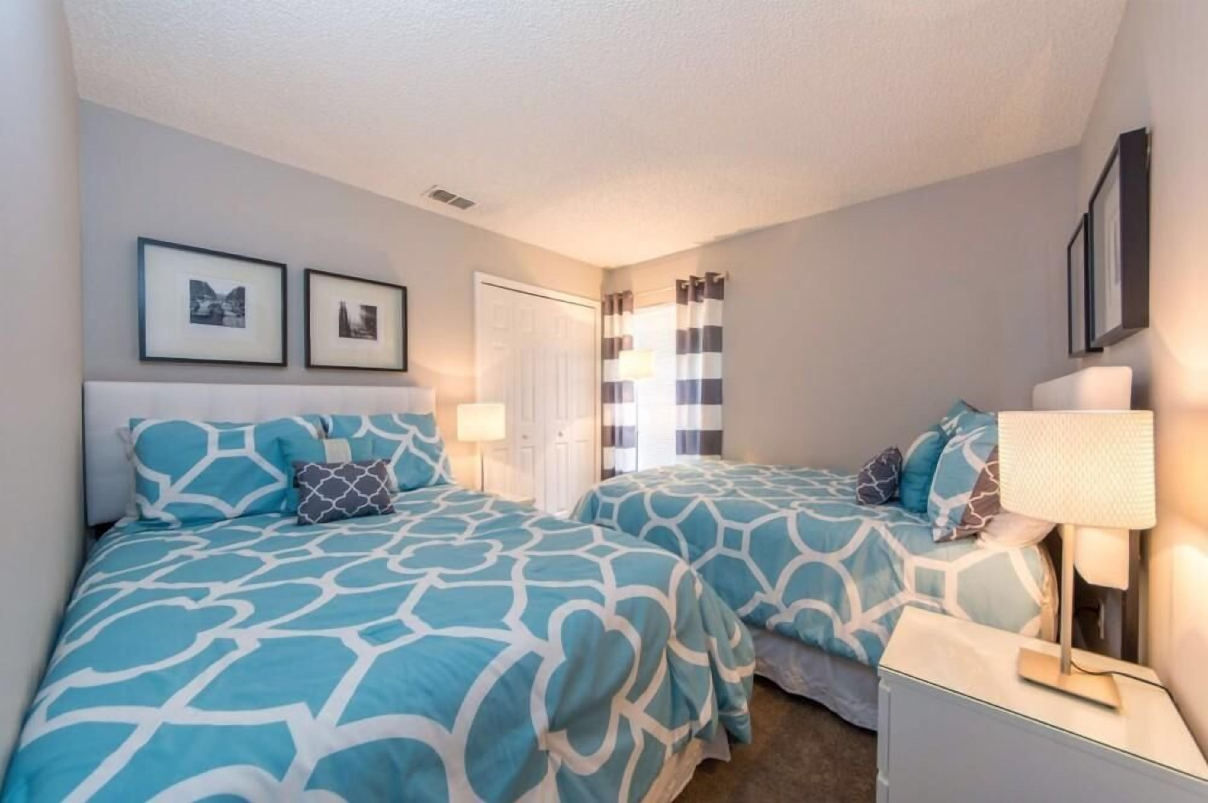 Un dormitorio con dos camas, cada una con ropa de cama azul y blanca a juego. Hay dos mesitas de noche con lámparas, una puerta y dos cuadros en la pared.