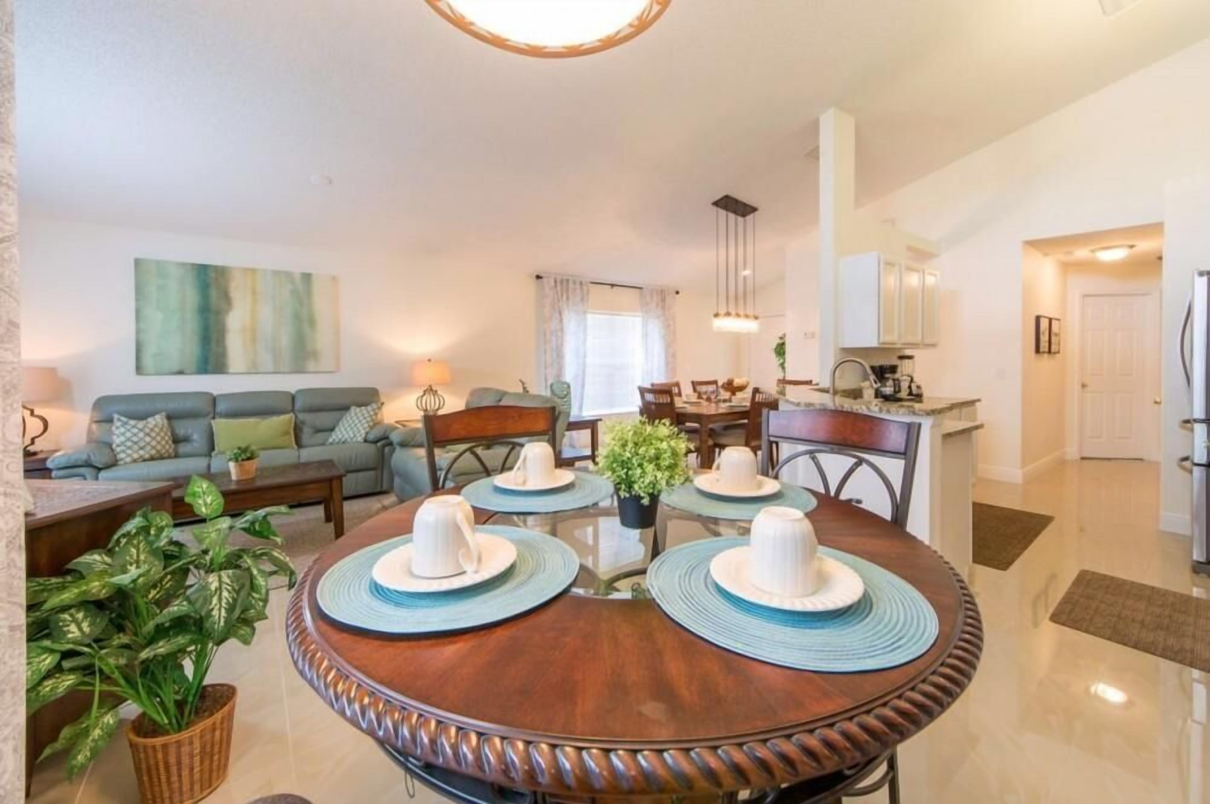 A modern living room with a dining table set for four, a sofa, and a kitchen in the background.