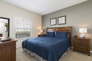 A bedroom with a large bed, two bedside tables with lamps, a dresser, and a window with blinds. - Villa Cristina - Lucaya Village (Kissimmee)
