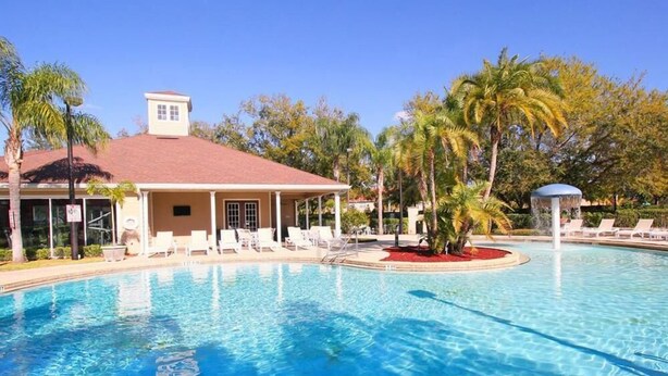 A swimming pool with lounge chairs and a covered area, surrounded by palm trees and a building with a red roof. - Villa Fonseca - Lucaya Village (Kissimmee)