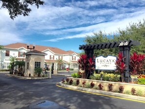 A gated entrance with a sign for Lucaya Village, featuring a clear sky and residential buildings in the background. - Villa Minnie - Lucaya Village (Kissimmee)
