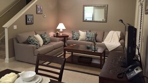Ein Wohnzimmer mit Sofa, Couchtisch und Schreibtisch mit Computer. Eine Lampe steht auf dem Couchtisch und ein Telefon auf dem Schreibtisch. Über dem Sofa ist ein Deckenventilator zu sehen.