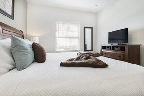 Una cama bien hecha con almohadas, un televisor sobre un soporte de madera y una ventana con persianas.