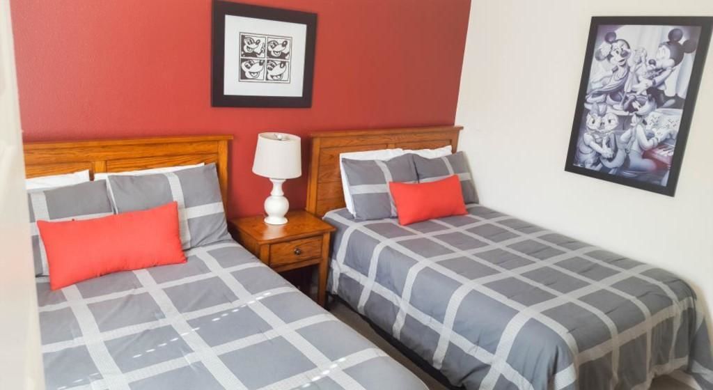 Quarto de hotel com duas camas, cada uma com colcha cinza e branca xadrez e almofadas vermelhas. Há uma mesa de cabeceira com luminária branca entre as camas.