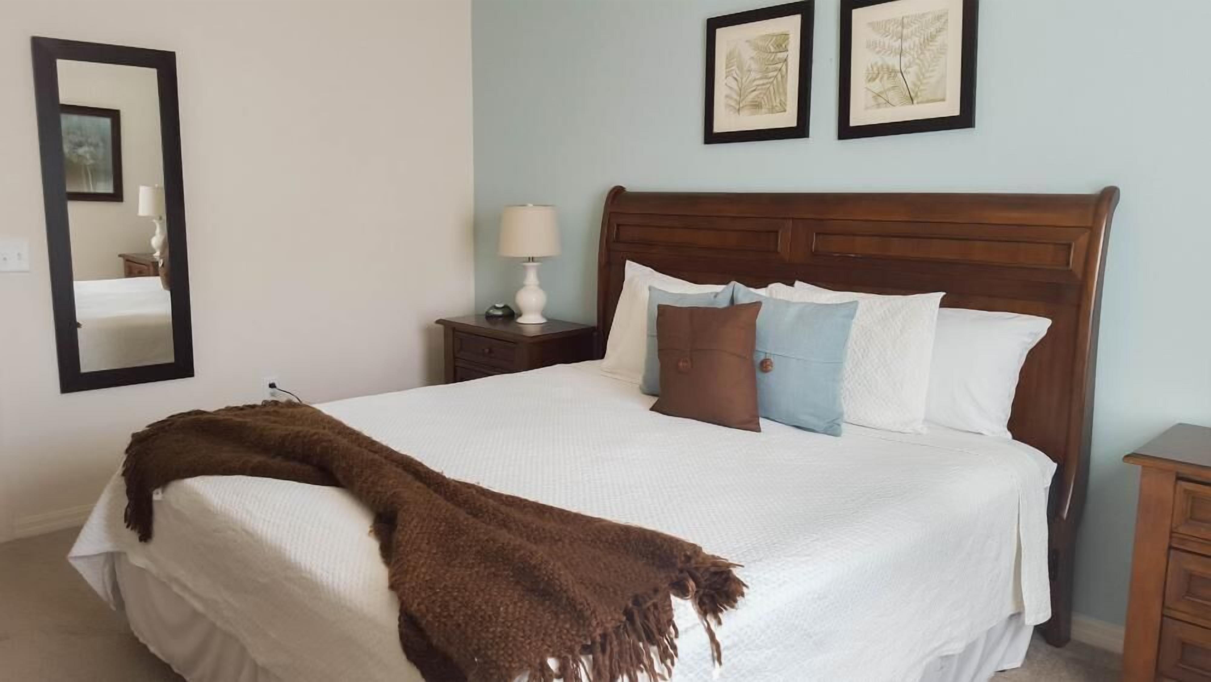 A neatly made bed with a brown throw blanket, a wooden headboard, and two framed botanical prints on the wall.