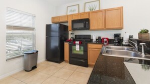 A compact kitchen with a refrigerator, microwave, oven, and sink. There is a trash bin and a window with blinds. - Villa Rodrigues - Encantada Resort (Kissimmee)