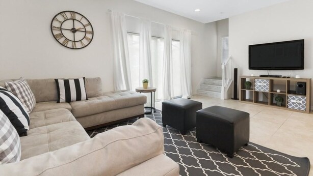 A modern living room with a sectional sofa, ottomans, a clock, a flat-screen TV, and a staircase leading to an upper level. - Villa Rodrigues - Encantada Resort (Kissimmee)