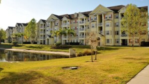 A row of multi-story residential buildings with balconies, a grassy area, and a small pond.