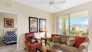 A living room with a bed, a red armchair, a beige sofa, a coffee table with a plant, and a large window with a view of buildings.