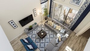 Una sala de estar moderna con un sofá gris, un sillón azul y una alfombra estampada. Un televisor de pantalla plana está montado en la pared, y hay una planta en maceta y una lámpara de pie.