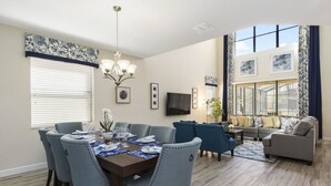 Un comedor con una mesa redonda, sillas azules y una araña. Hay una sala de estar con un sofá y un televisor.