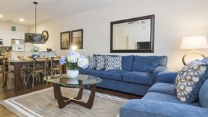 A living room with a blue sofa, a glass coffee table, and a dining area with chairs and a table. - Villa CVX- Lucaya Village (Kissimmee)