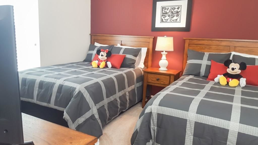 Une chambre d’hôtel avec deux lits, chacun agrémenté d’une peluche à l’effigie de Mickey Mouse, une table de chevet avec une lampe et un tableau encadré au mur.