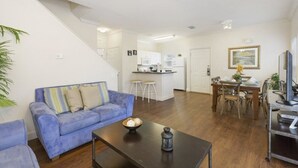Una sala de estar con un sofá azul, una mesa de centro con objetos decorativos, un comedor con una mesa y sillas de madera, y una cocina al fondo.