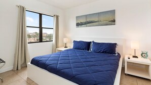 A neatly made bed with a blue quilt, a nightstand with a lamp, and a wall-mounted painting. - Villa Yamagi - The Enclave Hotel & Suites (Orlando)