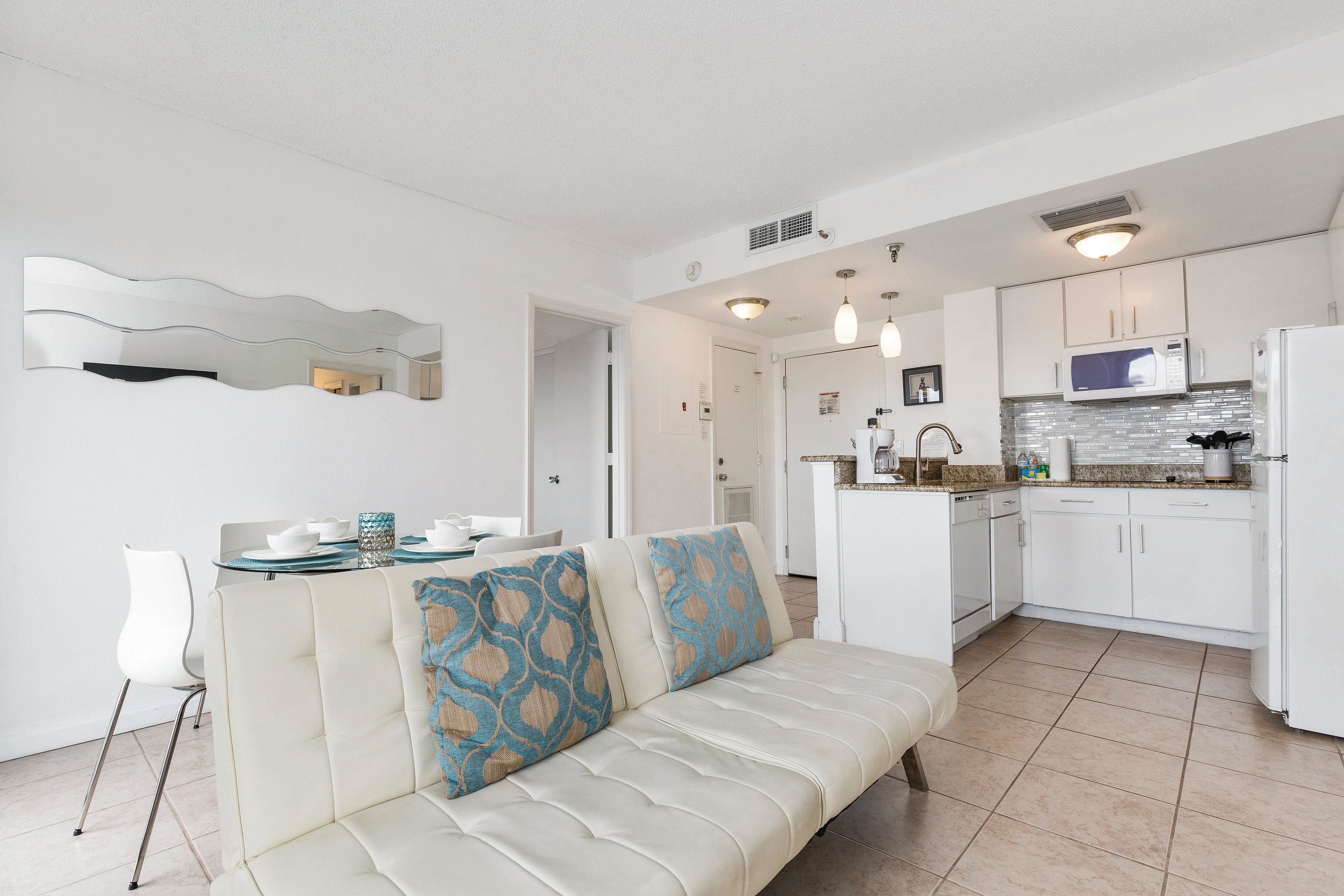 A modern kitchen with white cabinets, a microwave, and a refrigerator. There is a beige sofa with blue patterned cushions in the living area. - Villa Yamagi - The Enclave Hotel & Suites (Orlando)