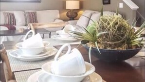 Uma sala de jantar com uma mesa posta para quatro pessoas, um arranjo de plantas decorativo e uma escada ao fundo.