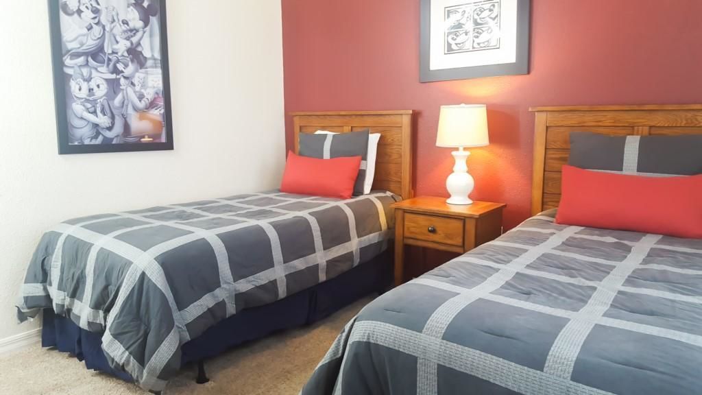 A hotel room with two beds, each with a grey and white checkered bedspread, red pillows, and a nightstand with a lamp between them.