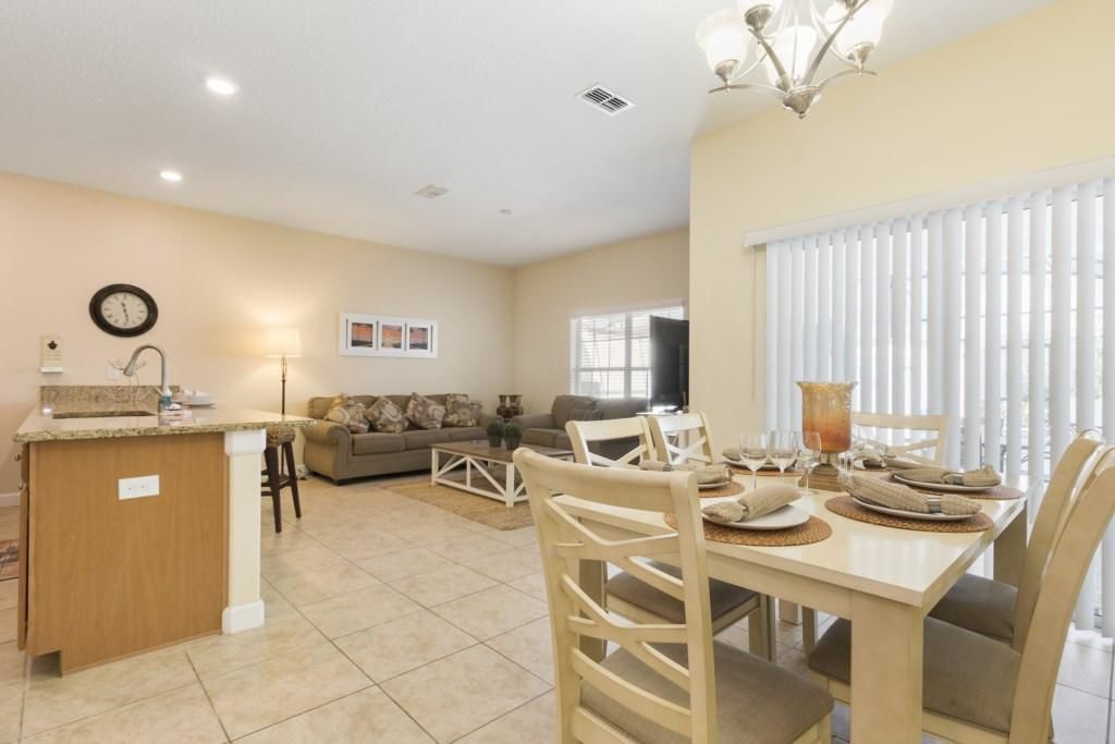 A modern kitchen with a central island, dining area with chairs and a table set for four, a sofa, and a TV in the background.