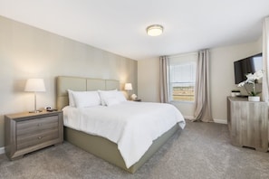 A bedroom with a bed, bedside tables, a nightstand, a dresser, a window with curtains, and a wall-mounted TV.