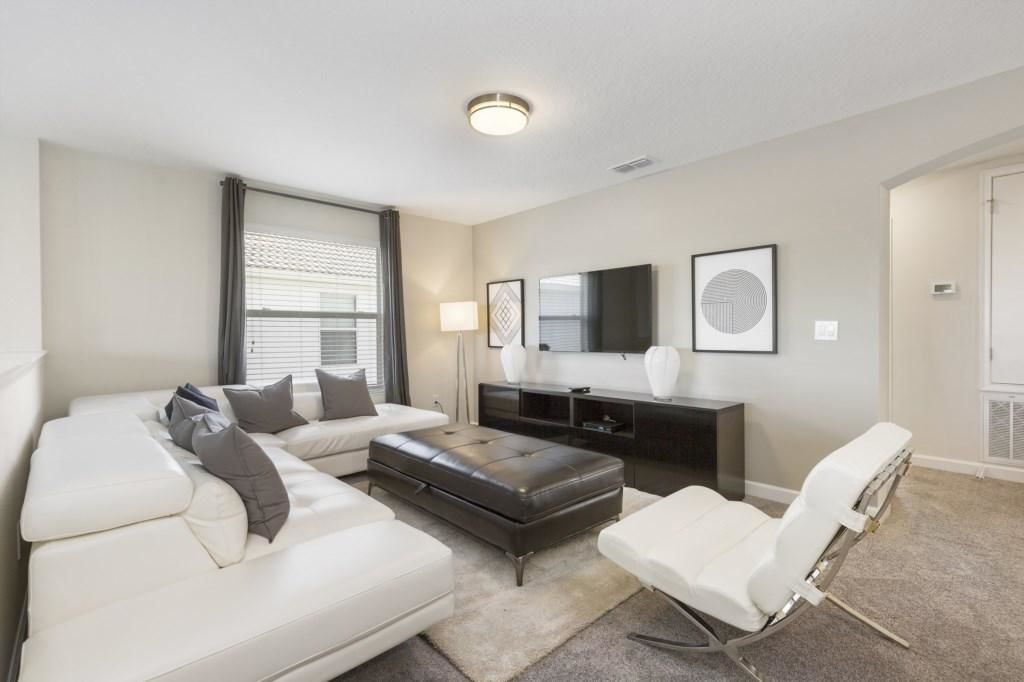 A modern living room with a sectional sofa, a white armchair, a leather ottoman, a flat-screen TV mounted on the wall, and a small side table with decorative vases.