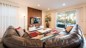 A modern living room with a leather sofa, a flat-screen TV, a coffee table, and a large window with a view of a swimming pool and buildings. - Villa Veni Vici - Encore Resort at Reunion (Kissimmee)