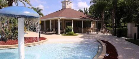 A pool area with a fountain, a covered seating area, and a building with a red roof.