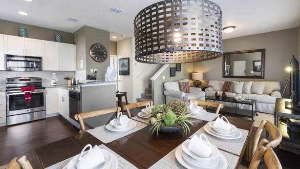 A modern kitchen with stainless steel appliances, a dining area with a wooden table set for four, and a living room with a sofa and a large mirror. - Villa Gusta - Lucaya Village (Kissimmee)