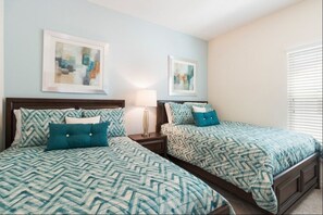 Quarto com duas camas, cada uma com roupa de cama estampada e almofadas combinando. Há quadros nas paredes, um abajur e uma janela com persianas.