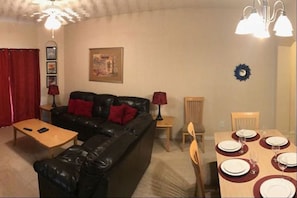 A living room with a black leather sofa, a wooden coffee table, a dining table set for four, and a red curtain.
