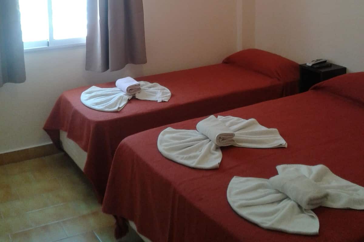 Deux lits simples avec du linge de lit rouge, une petite table avec des serviettes, une table de chevet et une fenêtre avec des rideaux.