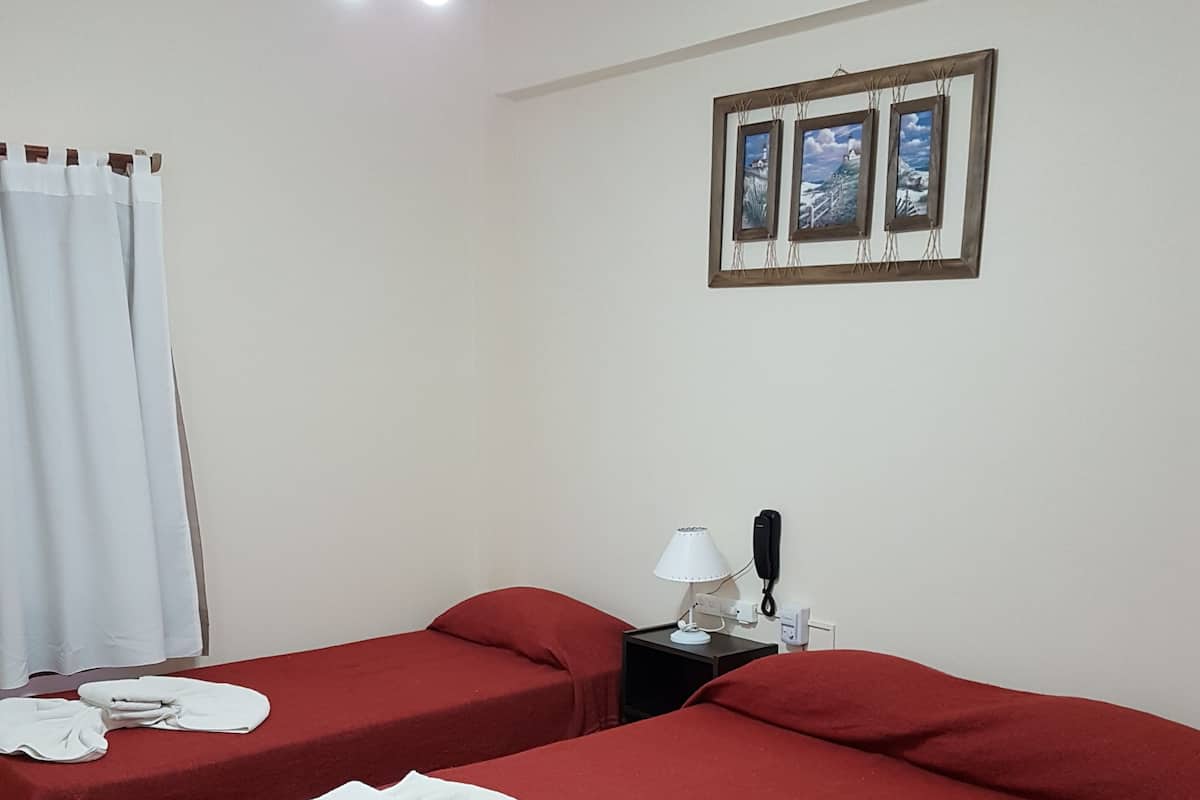 Une chambre d’hôtel avec deux lits, chacun recouvert d’une couverture rouge et agrémenté de serviettes blanches. On y trouve un ventilateur de plafond, une table de chevet avec une lampe et un tableau encadré au mur.