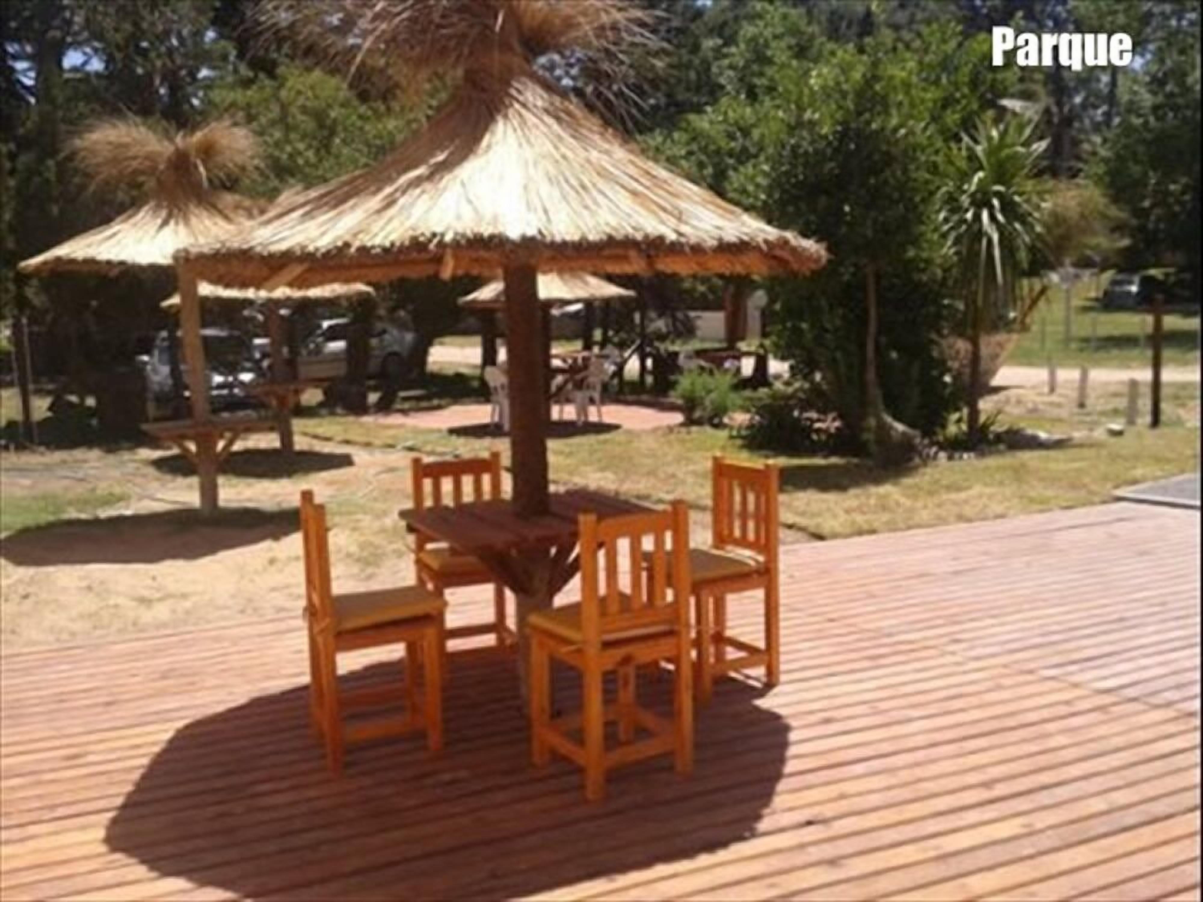 An outdoor wooden deck with a thatched-roof table and chairs, surrounded by greenery.