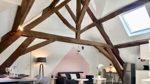 Een loft-achtige woonruimte met zichtbare houten balken, dakraam, eethoek met tafel en stoelen, een bank en een keuken.