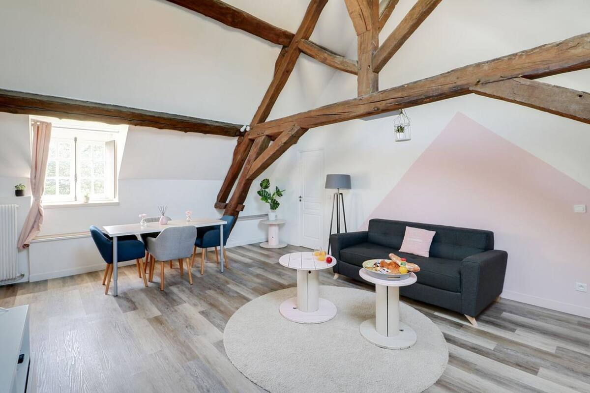A modern living room with exposed wooden beams, a dining area with blue chairs, a grey sofa, and a round coffee table.
