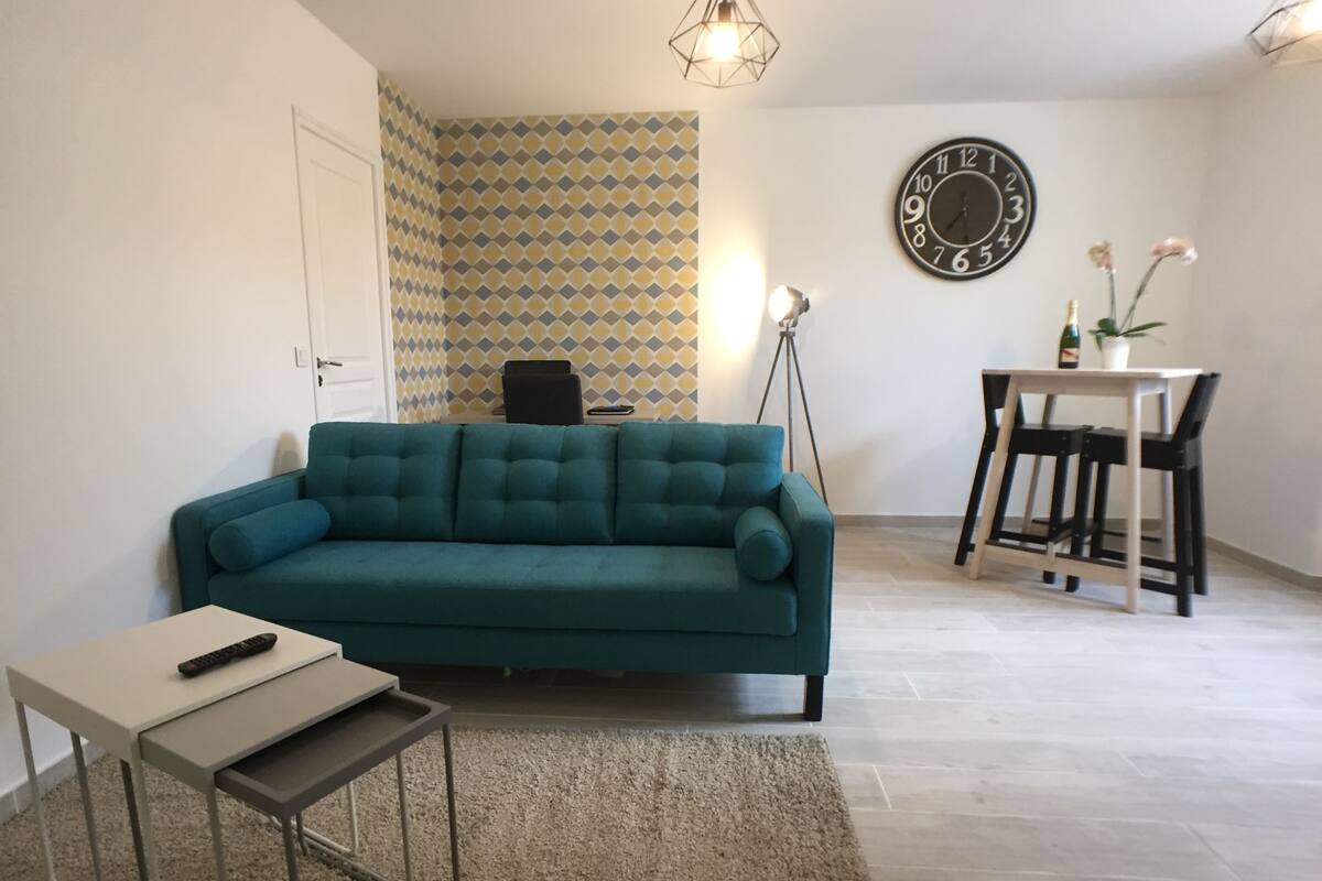 A living room with a teal sofa, a small table, a floor lamp, and a dining area with a table and chairs.