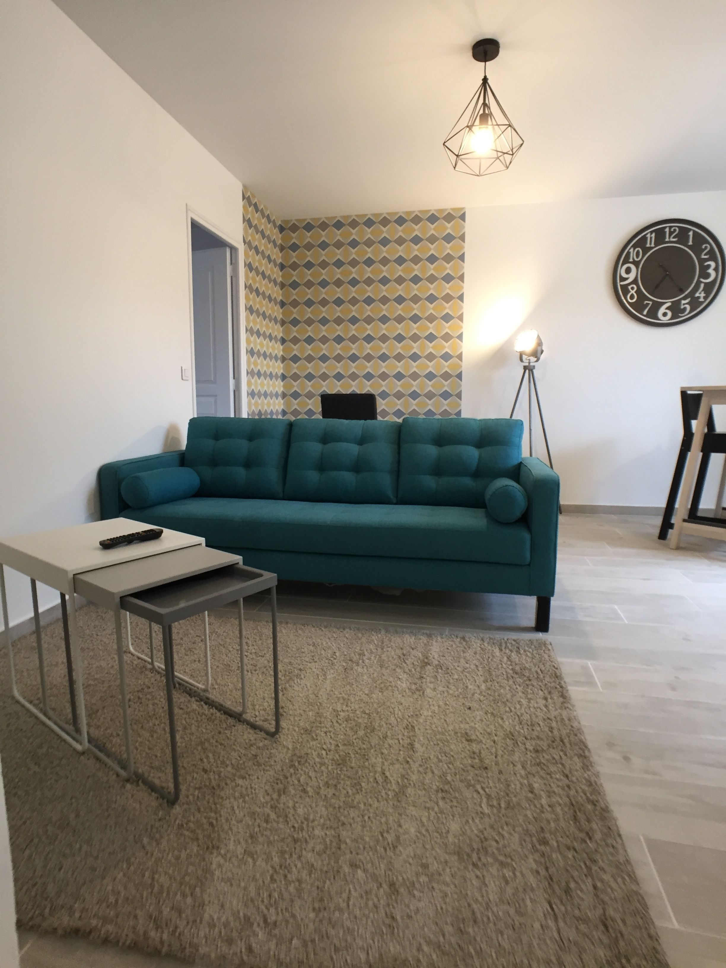 A living room with a teal sofa, a small side table, a patterned wall, a clock, and a floor lamp.