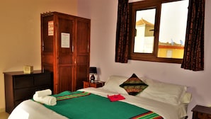 A hotel room with a bed, a wooden wardrobe, a nightstand, a window with curtains, and a view of buildings outside. - Auberge Etoile Mirleft (Mirleft)
