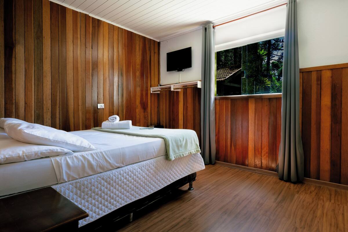 Ein Schlafzimmer mit einer holzverkleideten Wand, einem Bett mit weißer Bettwäsche, einem flachbildfernseher, der über dem Bett angebracht ist, und einem Fenster mit Vorhängen.