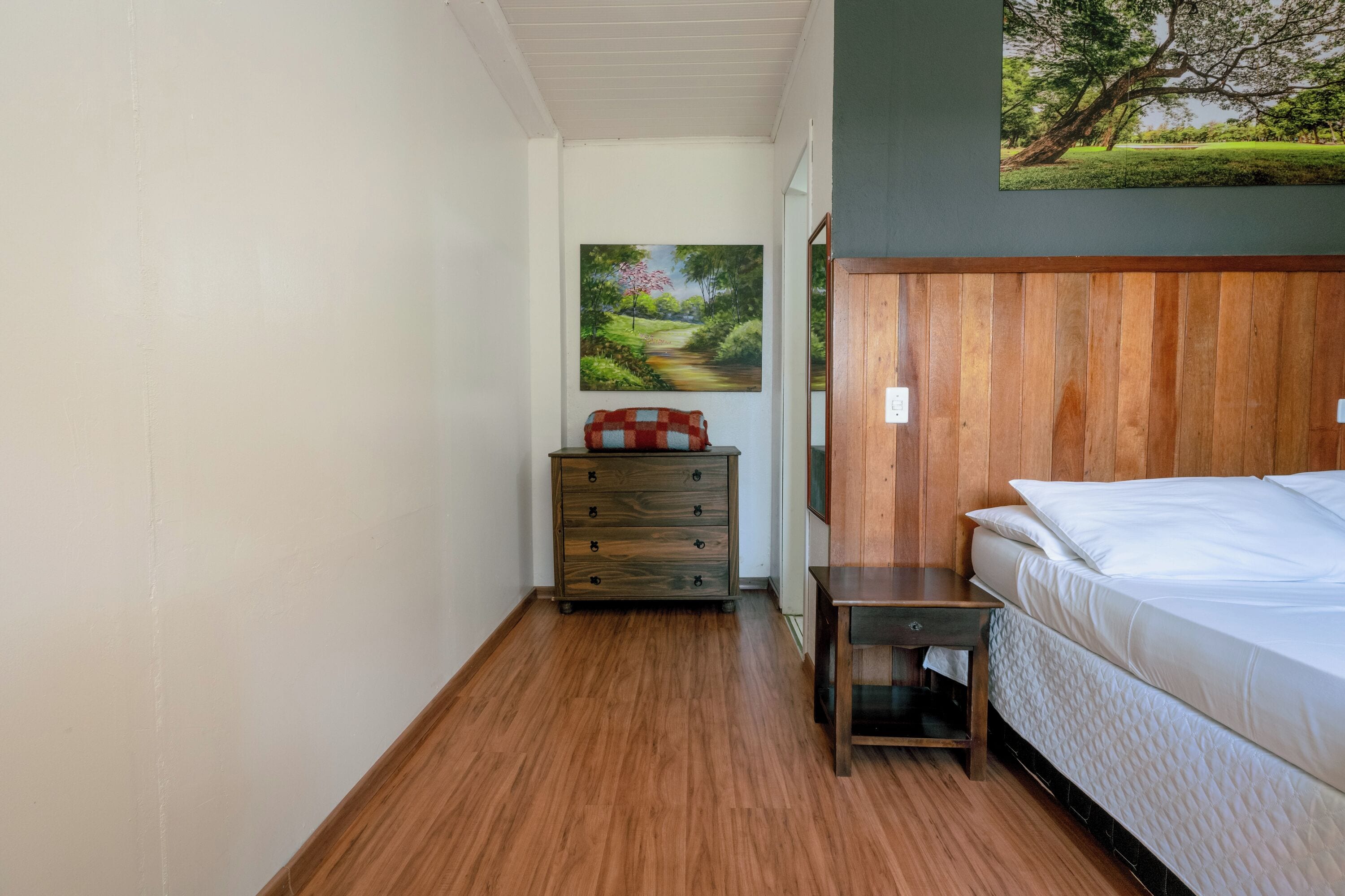 A bedroom with a wooden headboard, a bed, a nightstand, a dresser, and a painting of a landscape on the wall.