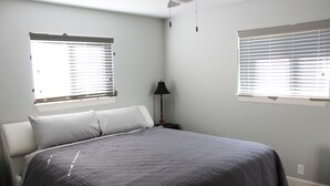 Une chambre à coucher avec un lit, deux fenêtres équipées de stores, une table de chevet avec une lampe et un sol recouvert d’une moquette.