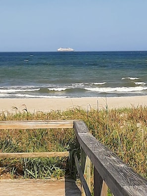 En träpromenad som leder till en strand med vågor och ett fartyg i fjärran.