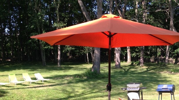 A wooden deck with a glass table and chairs, an orange umbrella, and a barbecue grill.