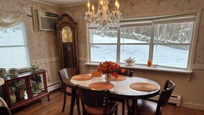 A dining room with a round table, chairs, a chandelier, a grandfather clock, and a window with a view of a snowy landscape.